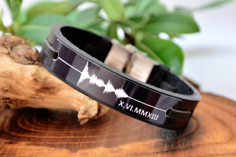 Puede incluir: Pulsera de cuero negro con cierre plateado. La pulsera presenta un dise&ntilde;o de onda de sonido blanco y la fecha en n&uacute;meros romanos X.VI.MMXIII. La pulsera se muestra sobre una superficie de madera con follaje verde en el fondo.