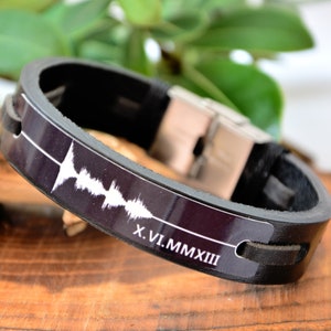 Puede incluir: Pulsera de cuero negro con cierre de acero inoxidable. La pulsera presenta un dise&ntilde;o de onda de sonido blanco y la fecha en n&uacute;meros romanos X.VI.MMXIII. La pulsera se muestra sobre una superficie de madera.