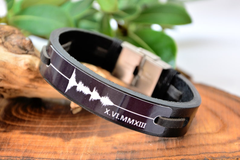 Puede incluir: Pulsera de cuero negro con cierre de acero inoxidable. La pulsera presenta un dise&ntilde;o de onda de sonido blanco y la fecha en n&uacute;meros romanos X.VI.MMXIII. La pulsera se muestra sobre una superficie de madera.