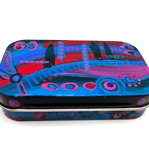 May include: A small, rectangular tin with a colorful abstract design. The design features swirls of blue, pink, red, and black paint with white dots and circles.