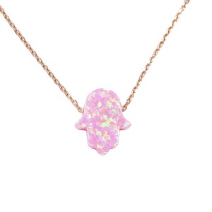 May include: A delicate gold chain necklace with a pink opal hamsa hand charm. The hamsa hand is a symbol of protection and good luck.