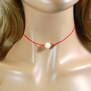May include: A delicate red cord necklace featuring a single, lustrous white pearl. The pearl is accented with small gold-colored beads. This minimalist necklace is a simple yet elegant piece of jewelry.