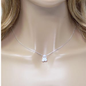 May include: A silver necklace featuring a small, white ghost pendant. The ghost has two eye holes and a curved bottom. The necklace is displayed on a mannequin.