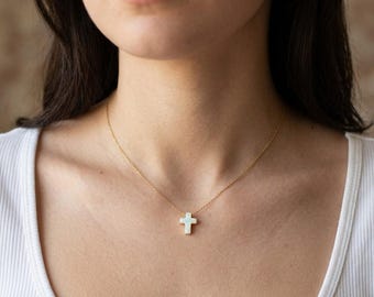 May include: A gold-colored necklace with a small, white cross pendant. The cross is centered on the chest, and the chain is delicate. The necklace is worn against a white top.