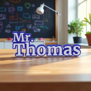 May include: A blue and white desk nameplate that reads "Mr. Thomas". The nameplate is in a bold, blocky font and sits on a wooden surface. The background includes a chalkboard and a desk lamp.