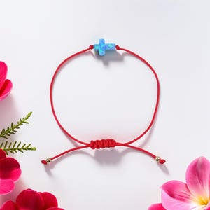 May include: A red string bracelet with a blue opal cross charm and gold beads. The bracelet is adjustable and can be tied to fit any wrist.