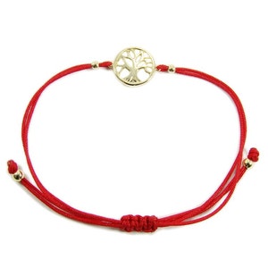 May include: A red string bracelet featuring a gold-colored tree of life charm. The charm is circular with a detailed tree design. The bracelet has adjustable knots and small gold-colored beads.