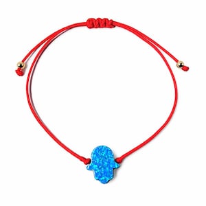 May include: Red string bracelet featuring a blue Hamsa hand charm. The adjustable bracelet has gold-colored beads at the ends and a sliding knot closure. The Hamsa hand is a vibrant blue with a textured appearance.