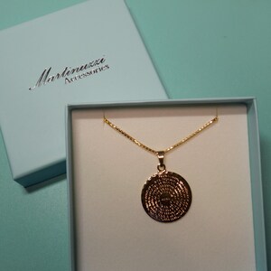 May include: A gold necklace with a round pendant featuring a detailed design. The necklace is displayed in a white box with a blue lid. The lid has the text "Martinuzzi Accessories" printed on it.