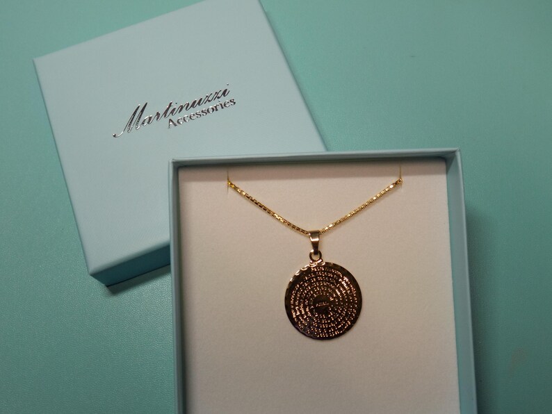 May include: A gold necklace with a round pendant featuring a detailed design. The necklace is displayed in a white box with a blue lid. The lid has the text "Martinuzzi Accessories" printed on it.
