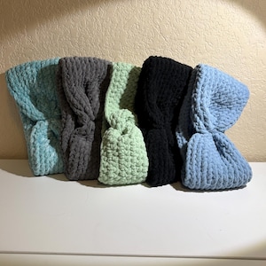 May include: Five handmade, knitted headbands in various colors: teal, gray, light green, black, and light blue. Each headband is made of a soft, textured yarn and has a twisted knot design. The headbands are displayed on a white surface.
