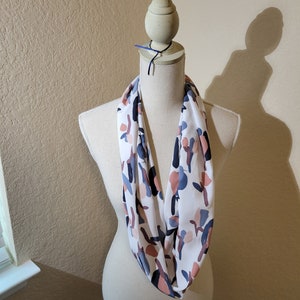 May include: A white infinity scarf with a colorful abstract print featuring shades of blue, pink, and brown.