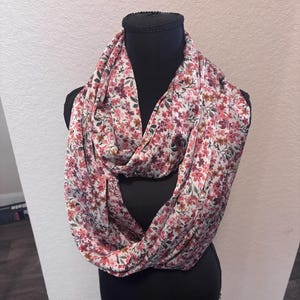 May include: An infinity scarf featuring a white base fabric with a dense, repeating floral pattern in shades of pink, red, and muted green. The lightweight fabric is draped in multiple loops around a black display form, creating a voluminous look.