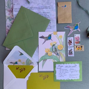 May include: A collection of stationery items, including envelopes, cards, and stamps, arranged on a pale green surface. The stationery features hummingbird illustrations and floral patterns. The color palette includes green, yellow, and floral patterns.
