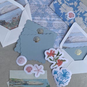 May include: A stationery set featuring light blue envelopes with floral patterns, scalloped-edge note cards with gold foil seashell accents, a letter with printed text, and various die-cut stickers depicting seashells, a starfish, and flowers.