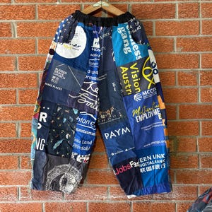 Puede incluir: Pantalones de pierna ancha confeccionados con un mosaico de varios paneles de tela azul, negra y blanca con diversos logotipos y textos impresos, incluidos Blessed Grace Church, PAYM y Jobs Fashion Link, con cintura elástica negra.