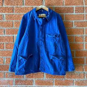 May include: A bright blue work jacket featuring a classic collar, a button-up front, and three large patch pockets. The garment includes a label at the inner neckline with the text HELRO. The jacket is constructed with long sleeves and a straight hem.