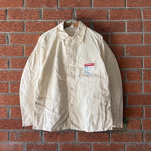 May include: A long-sleeved off-white work jacket featuring a classic collar, a button-down front, and two large lower patch pockets. A rectangular label with the red and blue text Pioneer is stitched onto the upper left chest pocket area.