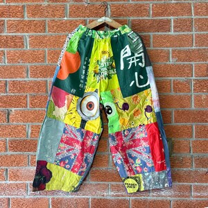 May include: Wide-leg trousers featuring a patchwork design of various colourful fabrics, including a Union Jack pattern, a cartoon eye graphic, and Japanese text characters. The trousers have an elastic waistband and are displayed on a wooden hanger.