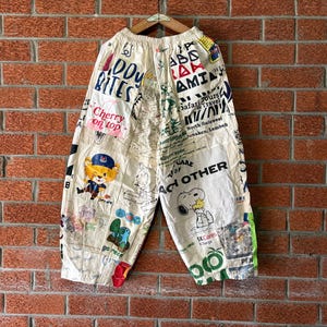 Könnte beinhalten: Cremefarbene Hose mit weitem Bein und einer Collage aus verschiedenen grafischen Drucken, darunter ein Cartoon-Bär, Snoopy sowie Schriftzüge wie "Diddy Bites", "Cherry on top", "Care of each other" und "Go Green", mit elastischem Bund und lockerer Passform.