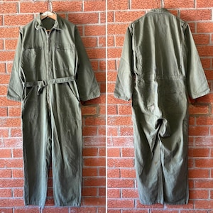 May include: An olive green utility jumpsuit featuring a collared neckline, a full-length front zip, long sleeves, and two chest pockets. The garment includes a matching fabric belt at the waist and two side pockets, shown from both front and back views.
