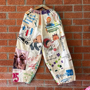 May include: Off-white wide-leg trousers featuring a collage of colourful graphic prints, including cartoon characters, a brown dog, text snippets like 'a big help to little minds', 'The Body Shop', and 'PychNed', with an elasticated waistband.