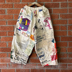 May include: Off-white loose-fitting pants featuring an all-over collage of various printed graphics, logos, and text including 'Gorged', 'Student Housing', and a Volvo emblem. The pants have an elasticized waistband and a wide-leg silhouette.