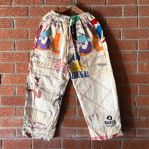 May include: Off-white pants featuring an all-over collage print of various colorful graphics, illustrations, and text including "Bared Footwear," "Libraries," and "Love." The pants have an elasticized waistband and a loose, wide-leg silhouette.