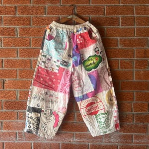 May include: Wide-leg pants featuring a patchwork design with various colorful graphic prints, including a unicorn, text reading "Stay united" and "Sparkgirl," and abstract patterns on a light-colored fabric base with an elasticized waistband.