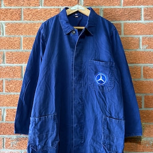 May include: A long-sleeved navy blue work coat featuring a classic collar, a button-down front, and two large lower pockets. A circular patch with a white Mercedes-Benz three-pointed star logo is stitched onto the left chest pocket area.