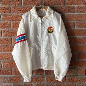 Può includere: Giacca vintage color crema con zip e colletto. La manica sinistra presenta strisce blu e rosse. Una toppa circolare con una scena di montagna è sul petto. La giacca è appesa a un muro di mattoni.