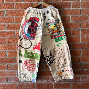 May include: Off-white pants featuring a collage of various graphic prints, including a Spider-Man illustration, a Community Potluck fish graphic, and assorted text elements like Good Morning Crer and Hogwarts. The pants have an elastic waistband.