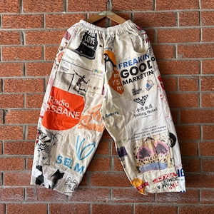 May include: Off-white wide-leg trousers featuring a patchwork design of various graphic prints, logos, and text including Radio Brisbane, Jinling Hotel, and Freakin Good Marketing. The trousers have an elastic waistband and are displayed on a wooden hanger.