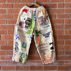 Könnte beinhalten: Cremefarbene Hose mit einem durchgehenden Collage-Druck aus verschiedenen bunten Logos, Texten und grafischen Illustrationen. Die Hose hat einen elastischen Bund und eine weite Beinform und zeigt verschiedene Markennamen und Slogans.