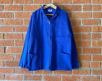 May include: A bright blue long-sleeve work jacket featuring a full-length front zipper, a pointed collar, one chest pocket, and two lower front pockets. The garment is displayed on a wooden hanger against a red brick wall background.