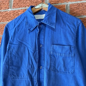 May include: A bright blue long-sleeve work shirt featuring a pointed collar, a front button-down closure with silver-toned metal buttons, and a single chest pocket on the left side. The garment is displayed on a wooden hanger against a brick wall.