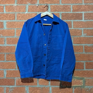 May include: A vibrant blue long-sleeve work jacket featuring a classic collar, a front button-down closure with four visible dark buttons, and two large patch pockets on the lower front. The jacket is displayed on a wooden hanger.