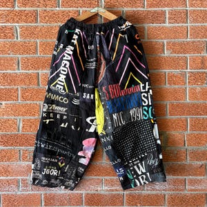 以下が含まれることがあります： NYC 1991、Army、New Yorkなどの様々なカラフルなグラフィックプリント、テキスト、ロゴがパッチワーク状にデザインされた黒のワイドパンツ。ウエストはゴム仕様で、木製のハンガーに掛けられた状態で表示されています。