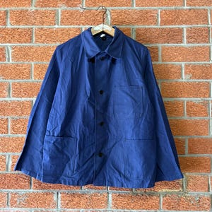 May include: A solid blue long-sleeve jacket featuring a classic collar, a front button closure, one chest pocket, and two lower patch pockets. The garment is constructed with a structured fabric and is displayed hanging on a wooden hanger.