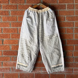 May include: Wide-leg pants featuring a patchwork design with a mix of white, light gray, and black grid patterns. The garment has an elasticized waistband, side pockets, and decorative horizontal stitching details near the bottom hemline.