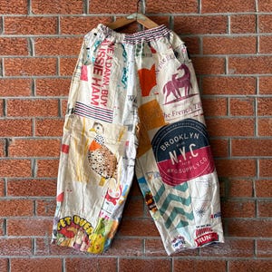 Könnte beinhalten: Cremefarbene Hose mit weitem Bein und einer Collage aus bunten Grafikdrucken, darunter ein Vogel, ein Pferd und der Text "BROOKLYN N.Y.C. MFG. SUPPLY CO." sowie "WE A DAMN, BUY AUSSIE HAM." Die Hose hat einen elastischen Bund und Seitentaschen.