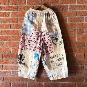 Könnte beinhalten: Weit geschnittene Hose im Patchwork-Design mit verschiedenen Mustern, darunter blaues Vichy-Karo, rosa Grafikdrucke, eine Katzenkopf-Illustration sowie der Text "WHAT'S IN YOUR BAG" und "Element". Die Hose hat einen elastischen Bund.
