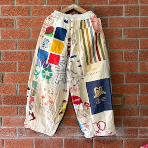 May include: Wide-leg trousers featuring a patchwork design of various printed fabrics in cream, blue, red, yellow, and green. The garment includes graphics of flamingos, a peace sign, a pretzel, and text including 60th Anniversary and Rues Sport.