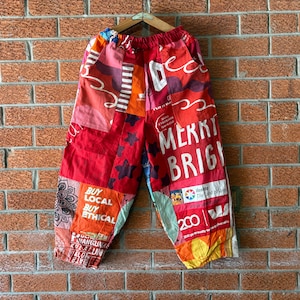 May include: Patchwork pants featuring a vibrant mix of red, orange, and multi-colored fabric panels. The design includes various printed text such as MERRY BRIGHT, BUY LOCAL BUY ETHICAL, and 200, along with star and mandala patterns. The pants have an elastic waistband.