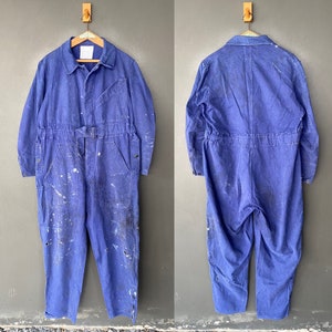Pode incluir: Um macacão de trabalho de brim azul com respingos de tinta branca. O macacão tem um fecho de correr frontal, dois bolsos grandes no peito e dois bolsos grandes nas pernas.