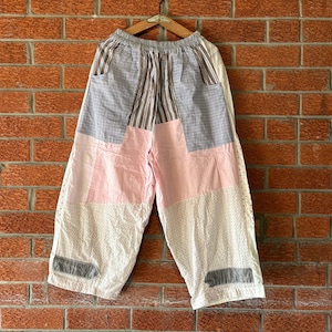 Può includere: Pantaloni a gamba larga con design patchwork caratterizzato da vari motivi di tessuto, tra cui righe verticali, pannelli in tinta unita rosa chiaro e tessuto bianco con piccoli punti scuri. I pantaloni hanno un elastico in vita e due toppe grigie.
