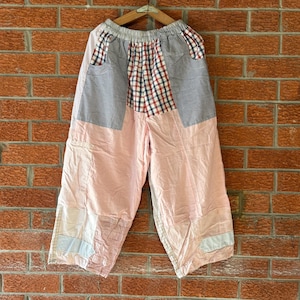 May include: Wide-leg pants featuring a patchwork design with light pink fabric, a red and blue plaid pattern, and small blue and white checkered sections. The pants have an elastic waistband and are displayed on a wooden hanger.
