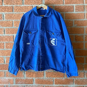 Può includere: Giacca blu brillante con due tasche sul petto e chiusura con bottoni. La giacca presenta un logo bianco sul davanti e una piccola etichetta sulla tasca sinistra. Appesa a una gruccia di legno contro un muro di mattoni.