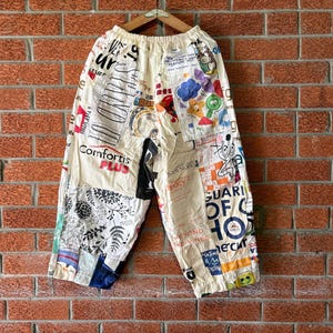 Puede incluir: Pantalones anchos de color blanco roto con un diseño de patchwork de varios tejidos estampados, incluyendo textos como Comfortis Plus, motivos florales e ilustraciones gráficas coloridas. Los pantalones tienen una cintura elástica y un corte holgado.