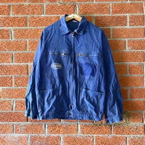 May include: A blue long-sleeve work jacket featuring a full-length front zip, a pointed collar, and two chest pockets with zips. The right chest pocket displays an oval patch with the text ARIS, while the left pocket has a flap.
